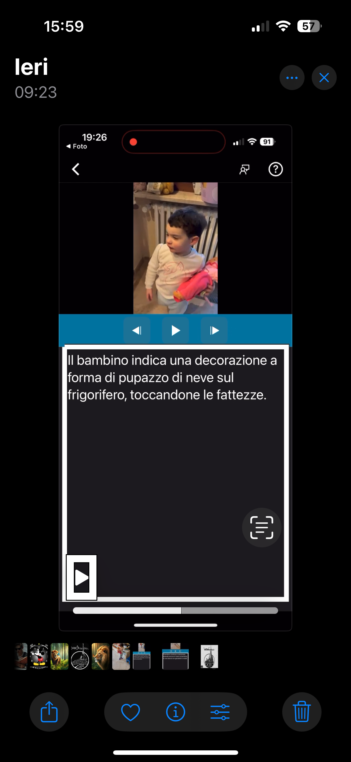 Nell'immagine uno screenshot di un video che nella parte superiore dello schermo, mostra un bambino in piedi in una cucina. Il bambino indossa una maglietta chiara e tiene in mano un oggetto colorato, forse una bambola. Sotto il video, c'è il testo dell’audiodescrizione: Il bambino indica una decorazione a forma di pupazzo di neve sul frigorifero, toccandone le fattezze. 